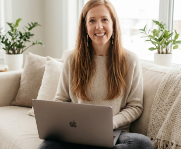 woman in her 40s sitting on a couch with a laptop on her lap smiling at the camera